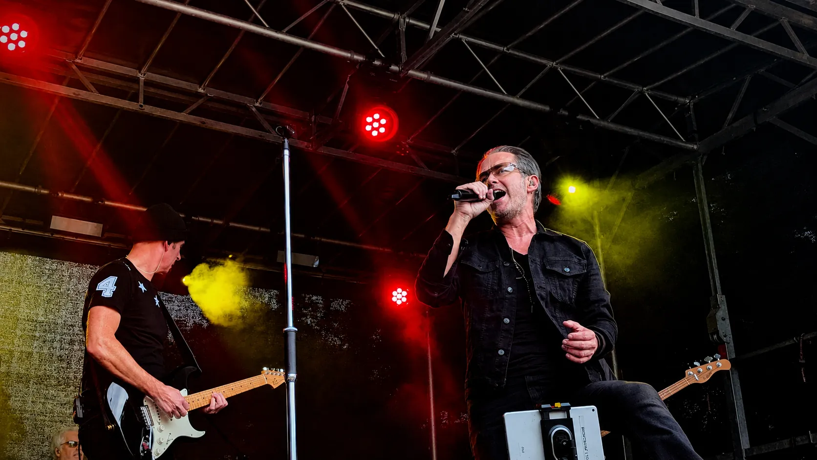 U2.V2 frontman singing with a foot up on the monitor, guitarist alongside, under red and yellow lights at Dumfries